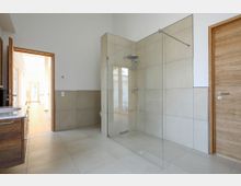 Modernes Badezimmer mit großer, bodenebener Dusche, umgeben von einer Glaswand. Die Wände und der Boden sind mit beigen Fliesen verkleidet, und Türen aus Holz führen in angrenzende Bereiche.