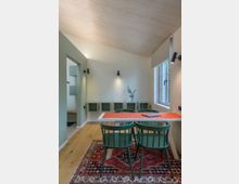 Kleiner, modern eingerichteter Raum mit Holzdecke und hellem Holzboden. In der Mitte steht ein weißer Tisch mit grün gestrichenen Holzstühlen, darüber hängt eine orangefarbene Tischdecke; darunter liegt ein farbenfroher Teppich mit orientalischem Muster. An der Wand befinden sich grüne Wandregale und ein Fenster mit beigeamer Jalousie.