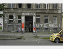 Fassade eines alten, grauen Mehrfamilienhauses mit sichtbarem Verfall und zahlreichen Graffitis sowie Plakaten an den Wänden und der Eingangstür. Vor dem Gebäude stehen ein Fahrrad, mehrere Poller und ein kleines gelbes Auto, auf dessen Seite die Aufschrift einer Arztpraxis zu sehen ist.