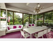 Moderner Wintergarten mit großen, bodentiefen Glasfenstern, die Blick auf einen grünen Garten mit Büschen und Bäumen bieten. Der Innenraum ist mit weißen Möbeln und pinken Akzentkissen sowie einem weißen Esstisch mit Stühlen eingerichtet.