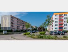 Mehrstöckige Wohnblöcke in einer Wohnsiedlung, links ein rosa Gebäude mit Balkonen, rechts ein weißes Gebäude mit bunten Balkonen in Rot und Gelb. Dazwischen befinden sich ein Parkplatz mit geparkten Autos, Gehwege und einige Bäume. Ein Straßenschild mit der Aufschrift "Heinrich-Heine-Str." ist sichtbar.