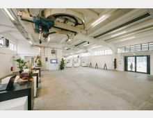 Industriehalle mit weiß gestrichenen Wänden, Oberlichtern und einem Betonboden. Der Raum ist teils mit Pflanzen, Ausstellungstischen und modernen Installationen ausgestattet, im Hintergrund sind Exponate und ein großes Eingangsportal mit Glastüren sichtbar.