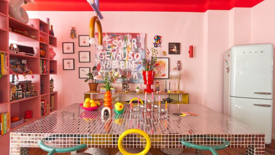 Bright, colorful kitchen-dining space with a glossy red ceiling, pink walls, and a large square mosaic-tiled table surrounded by modern chairs. Open shelving lines one wall, framed art and vases decorate the back wall, and a retro-style white refrigerator stands to the right.