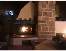 Gemütliches Wohnzimmer mit einem brennenden Kamin, umgeben von einer Verkleidung aus naturfarbenem Stein; im Vordergrund ein Tisch mit einer Dekoration, im Hintergrund eine Eingangstür mit beleuchtetem Kranz.