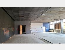 Großer, leerer Innenraum eines verlassenen Gebäudes mit abgehängter Decke, beschädigtem Boden und grauen Wänden mit Graffiti. Rechts verläuft eine Fensterfront mit Heizkörpern darunter, hinten befindet sich eine offene Tür zu einem dunkleren Nebenraum.