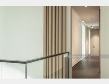 Minimalistischer Innenbereich mit hellem Flur, gläsernem Geländer mit Metallrahmen und einer dekorativen Holzwand mit vertikalen Paneelen. Der Boden ist aus dunklem Holz, und der Flur führt zu Türen mit glatten, weißen Oberflächen.