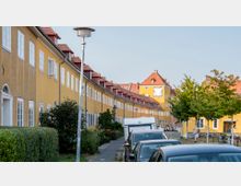 Straßenansicht in einem Wohngebiet mit gelben Reihenhäusern und roten Ziegeldächern, flankiert von Bäumen auf der rechten Seite. Im Hintergrund ist ein größeres Gebäude mit Uhr und hellem Fassadenbild sichtbar, während entlang der Straße Autos parken.