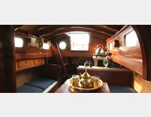 Holzverkleideter Innenraum eines Bootes mit abgerundeter Decke, kleinen Fenstern an den Seiten und einem runden Bullauge. Im Vordergrund ein Tisch mit Teegeschirr, im Hintergrund eine kleine Kochnische mit einem Wasserkocher und Stauraum.
