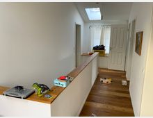 Ein heller Flur mit weiß gestrichenen Wänden und Holzboden. Links verläuft ein Geländer mit Ablagefläche, auf der Kinderspielzeug liegt; im Hintergrund ist eine Tür, ein Fenster mit Vorhang und ein Wäschekorb sichtbar.