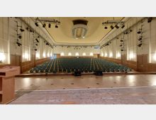 Großer Vortragssaal mit einer Bühne im Vordergrund, grünen Sitzreihen im Zuschauerraum und einer Decke mit einem ovalen, dekorativen Element. Die Wände sind beige und mit Holzpaneelen verkleidet, an denen Lampen und Bühnenscheinwerfer angebracht sind.