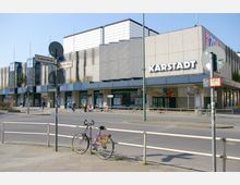 Außenansicht eines Karstadt-Warenhauses an einer Straßenkreuzung. Zu sehen sind ein modernistisches Gebäude mit grauer Fassade, große Schaufenster, Straßenschilder der Feuerbachstraße und Schloßstraße sowie ein geparktes Fahrrad im Vordergrund.