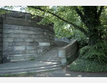 Steintreppe mit geschwungener Balustrade vor einer massiven Steinmauer, die teilweise mit Graffiti bedeckt ist, umgeben von dichtem Grün und Bäumen.