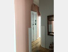 Ein Flur in einer Wohnung mit Altbau-Charakter, rosa gestrichenen Wänden und weißen Türrahmen. Der Boden besteht aus Holzdielen, an der Wand hängt ein Spiegel mit einem dekorativen Holzrahmen, und im Vordergrund steht ein geflochtener Korb.