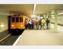 Eine U-Bahn-Station mit einer orangenen U-Bahn, auf der "Schlesisches Tor" angezeigt wird, steht an einem Bahnsteig. Eine Menschenmenge versammelt sich nahe der Bahn, und die Umgebung zeigt grüne Stützsäulen und eine leicht beleuchtete, unterirdische Atmosphäre.