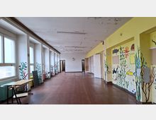 Langer, leerer Flur in einem heruntergekommenen Gebäude mit großen Fenstern auf der linken Seite und abblätternder Decke. An den gelben Wänden sind bunte Unterwasser-Wandmalereien mit Fischen und Pflanzen zu sehen; am Ende des Flurs führen offene Türen in weitere Räume.