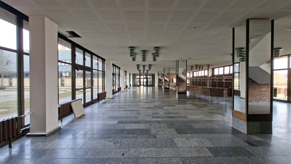Große, leere Eingangshalle oder Foyer mit grauem Steinboden, weißer Rasterdecke und Reihen von Deckenleuchten. Entlang der linken Seite verlaufen bodentiefe Fensterfronten, rechts stehen eine Garderoben-/Thekenzone und ein Treppenaufgang neben Ziegelwänden.