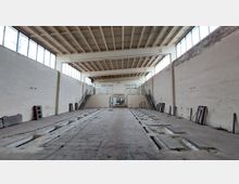 Ehemalige Fabrikhalle mit einer hohen Decke, rechteckigen Aussparungen im Boden und großen Fensterreihen entlang der Wände. An beiden Seiten führen Treppen zu erhöhten Bereichen, die in der Mitte von einer offenen Tür verbunden sind.