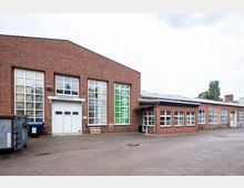Industrielles Backsteingebäude mit großen Fensterfronten und einem weißen Rolltor. Der Vorhof ist gepflastert, und einige Müllcontainer stehen seitlich des Gebäudes.