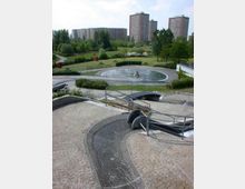 Eine städtische Parklandschaft mit Rasenflächen, Bäumen und Sitzbänken; im Vordergrund ein gepflasterter Bereich mit einem Wasserbecken und geschwungenen Rinnen. Im Hintergrund sind mehrstöckige Wohngebäude und weitere Grünflächen zu sehen.