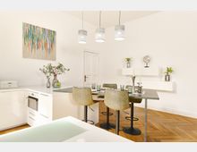 Moderne Küche und Essbereich mit hellen weißen Wänden, einem Holzboden und einer minimalistischen Einrichtung. Der Raum beinhaltet eine Kücheninsel mit Hockern, ein Essbereich mit vier Stühlen, hängenden Lampen, Wandregalen mit Dekoration sowie ein farbenfrohes abstraktes Gemälde an der Wand.