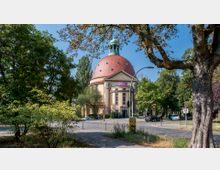 Straßenszene mit einer historischen Kuppelkirche, die durch ihre rote Kuppel und klassizistische Architektur mit Säulen und Rundfenstern auffällt, umgeben von Bäumen. Im Vordergrund sind ein gepflasterter Weg, zahlreiche Straßenschilder und geparkte Autos sichtbar.