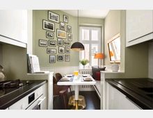 Kleine, helle Küche mit einer eingebauten Sitzecke um einen weißen Esstisch mit goldener Basis. Die Wände sind olivgrün gestrichen und mit einer Galerie aus gerahmten Schwarz-Weiß-Fotografien dekoriert; ein großes Fenster sowie ein schwarzer Pendelleuchter und dezente Einrichtungselemente wie eine Tischlampe und Kissen runden den Raum ab.