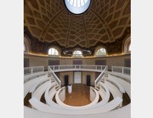 Innenraum eines historischen Hörsaals mit halbrunder, gestufter Sitzanordnung und weißen Balustraden. Die ovale Decke hat ein geometrisches Muster, eine große Kuppelöffnung und umlaufende Fenster mit dekorativen Malereien.