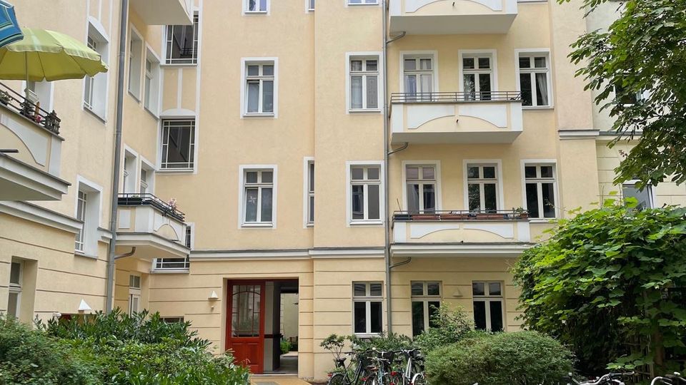 Innenhof eines mehrstöckigen Wohnhauses mit hellgelber Fassade, weißen Fensterrahmen und mehreren Balkonen sowie rotem Ziegeldach. Ein gepflasterter Weg führt zum Hauseingang, flankiert von Grünflächen und Fahrradständern mit abgestellten Fahrrädern.