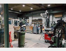 Werkstattinnenraum mit Industriemaschinen, Regalen voller Werkzeuge, Kisten und Kabeln. Metallische Oberflächen, eine Rohrlüftung und Beleuchtung an der Decke prägen den Raum.