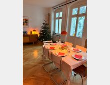 Helles Esszimmer mit Parkettboden, großem Esstisch mit weißer Tischdecke und gedeckten Plätzen sowie einer orangefarbenen Hängelampe darüber. Neben hohen Fensterfronten steht ein geschmückter Weihnachtsbaum; im Hintergrund ein Sideboard mit Tischlampe und Wandbild.