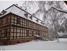 Fachwerkgebäude mit verschneiter Fassade und schneebedecktem Dach, umgeben von einer winterlichen Gartenlandschaft mit kahlen Bäumen und Sträuchern. Der Himmel ist grau und es liegt Schnee auf dem Boden.