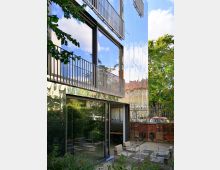 Moderne Gebäudefassade mit spiegelnder, metallischer Oberfläche und großen Glasfenstern, die Bäume und den Himmel reflektieren. Im Vordergrund ein kleiner Innenhof mit Sitzmöbeln und Pflanzenelementen, umgeben von einer Backsteinmauer.