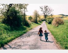 Zwei Kinder laufen auf einem schmalen, asphaltierten Weg durch eine ländliche Landschaft mit grünen Wiesen, blühenden Bäumen und einem gelb leuchtenden Rapsfeld im Hintergrund. Der Weg ist von Gras und Sträuchern gesäumt und führt in eine leicht hügelige Landschaft.