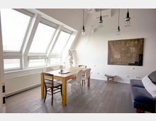 Minimalistisch eingerichtetes Esszimmer mit schrägen Dachfenstern, einem Holztisch, gemischten Stühlen und einer grauen Couch. Im Hintergrund hängen Lampen mit Drahtschirmen und ein großformatiges abstraktes Kunstwerk.