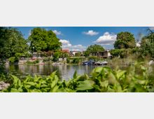 Ein ruhiges Gewässer mit Booten am Ufer, umgeben von dichter Vegetation und kleinen Gebäuden mit roten Dächern; im Hintergrund große Bäume und blauer Himmel mit weißen Wolken.