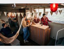Eine kleine Gruppe von Personen befindet sich auf einem Boot mit Blick auf das Wasser. Im Vordergrund steht eine Holztheke, dekoriert mit Obst und Getränken, während Wimpelketten die Umgebung schmücken; im Hintergrund sind Hängematten und ein gemütlicher Innenraum zu sehen.