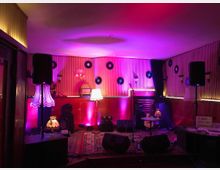 Ein kleiner Veranstaltungsraum mit einer Bühne, dekoriert im Retro-Stil mit Schallplatten an den Wänden, alten Lampen und Teppichen. Auf der Bühne stehen Mikrofone, Lautsprecher und ein Tisch mit einer Lampe, beleuchtet in violettem und warmem Licht.