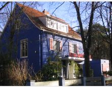 Blau gestrichenes Gebäude mit rotem Satteldach und weißen Fensterrahmen, das als Apotheke („Kur-Apotheke“) genutzt wird. Es ist von Bäumen und Sträuchern umgeben, die teilweise kahle Äste haben, und liegt in einer ruhigen, bewaldeten Umgebung.