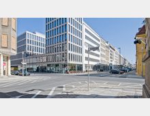 Straßenszene in einer Stadt mit modernen Bürogebäuden, gekennzeichnet durch helle Fassaden mit großen Fenstern. Im Vordergrund ist eine Kreuzung mit klar markierten Fahrbahnen und Straßenschildern, während parkende Autos und Fahrräder den Straßenrand säumen.