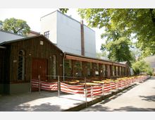 Außenbereich eines Restaurants mit Holzfassade und überdachter Terrasse; davor stehen gelbe Tische und rote Stühle, die durch rot-weiß gestreifte Absperrungen abgegrenzt sind. Im Hintergrund sind Bäume und ein größeres Gebäude mit Metallfassade zu sehen.