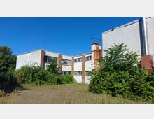 Ein leerstehendes, graues Gebäude mit rechteckigen Fenstern und sichtbaren Roststellen an einem Schornstein. Im Vordergrund wuchert dichter Bewuchs, und im Hintergrund ist ein klarer blauer Himmel sichtbar.