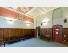 Innenraum eines historischen Gebäudes mit Holzvertäfelung an den Wänden, dekorativen Elementen und Sitzbänken. Die Decke ist kunstvoll mit Ornamenten gestaltet, und eine Uhr hängt an der Wand über der Vertäfelung.