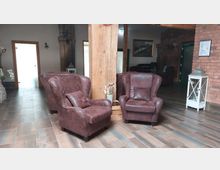 Gemütlicher Innenraum mit zwei braunen Ledersesseln und einem Ledersofa vor einem Holzpfosten. Der Raum hat braune Bodenfliesen, Backsteinwände, eine Laterne im weißen Holzrahmen und dekorative Pflanzen.