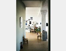 Ein heller Raum mit Holzdielen und weißen Wänden, geschmückt mit zahlreichen Kunstwerken und Fotos in verschiedenen Rahmen. Im Hintergrund stehen Pflanzen, ein mit bunten Textilien bedeckter Stuhl und ein kleiner Tisch, während im Vordergrund zwei kleine Holzstühle zu sehen sind.