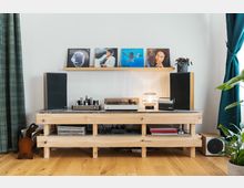 Holztisch mit Audio-Equipment, darunter ein Röhrenverstärker, ein Plattenspieler und Lautsprecher. Darüber an der Wand ein Regal mit Schallplatten, neben dem Tisch Pflanzen und ein Vorhang.