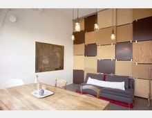 Raum mit modernem Design, bestehend aus einer hölzernen Tischgruppe im Vordergrund, einer grauen Couch mit weißem Kissen im Hintergrund und einer Wandverkleidung aus quadratischen Holz- und Farbpaneelen. Hängelampen mit sichtbaren Glühbirnen sowie ein abstraktes Kunstwerk ergänzen die Einrichtung.