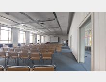 Ein Konferenzraum mit einer großen Anzahl an Stühlen, die in Reihen aufgestellt sind, und einem Projektorbildschirm an der Stirnseite. Der Raum hat hohe Fenster, die Tageslicht hereinlassen, sowie eine weiße Decke mit sichtbaren Beleuchtungs- und Kabelstrukturen.