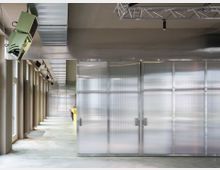 Moderner Innenraum mit einem breiten Korridor, transluzenten Wänden aus Polycarbonat und einer offenliegenden Decke mit Metallträgern und Lüftungskanälen. Links befinden sich hohe Fenster mit Heizkörpern darunter, und rechts ist ein gelber Tresen sichtbar.