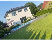 Modernes zweigeschossiges Wohnhaus mit grauem Satteldach und großen Fenstern, davor eine Terrasse mit Sonnenschirm und ein rechteckiger Pool. Im Vordergrund liegt ein großer, gepflegter Rasen, umgeben von Hecken und Bäumen in einem Wohngebiet.
