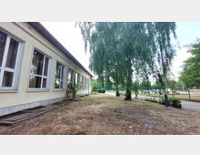 Ein einstöckiges Gebäude mit hellgelber Fassade und großen Fenstern, daneben ein Baum mit weit herabhängenden Zweigen. Im Hintergrund sind ein Weg, eine umzäunte Grünfläche und Parkbänke zu sehen.